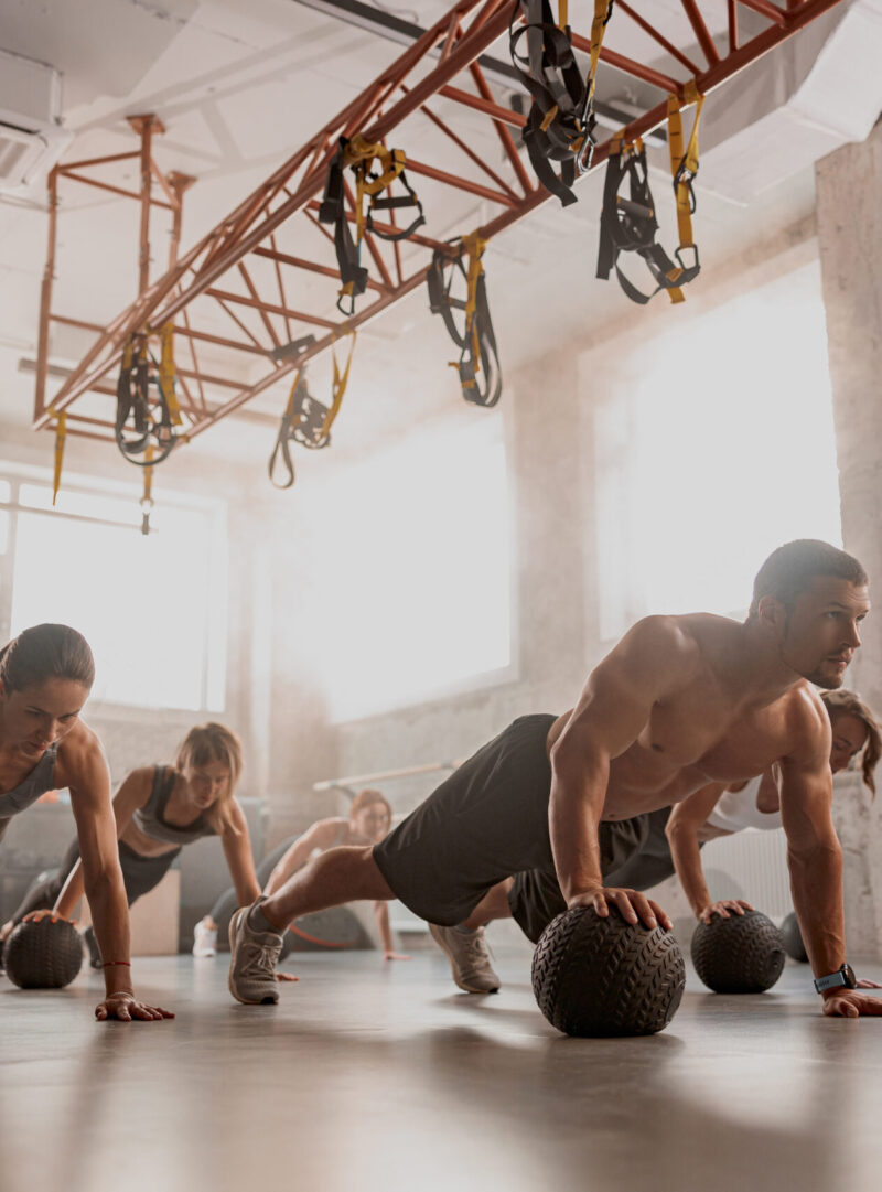 Handsome shirtless muscular male trainer and sporty women using exercise balls while having workout at industrial gym