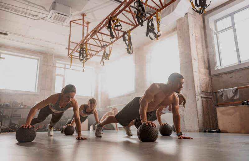 Handsome shirtless muscular male trainer and sporty women using exercise balls while having workout at industrial gym