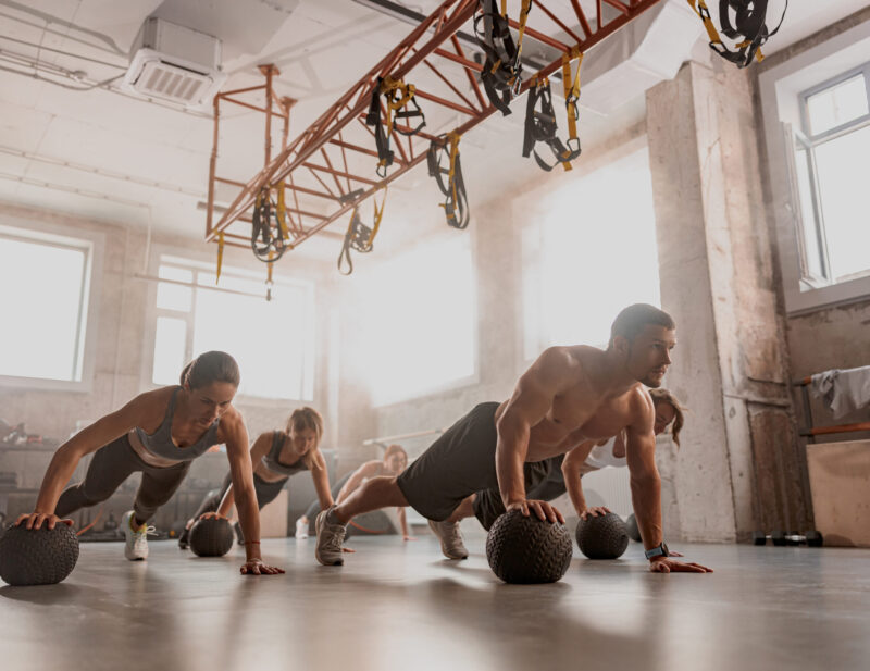 Team of young people training in gym club Handsome shirtless muscular male trainer and sporty women using exercise balls while having workout at industrial gym